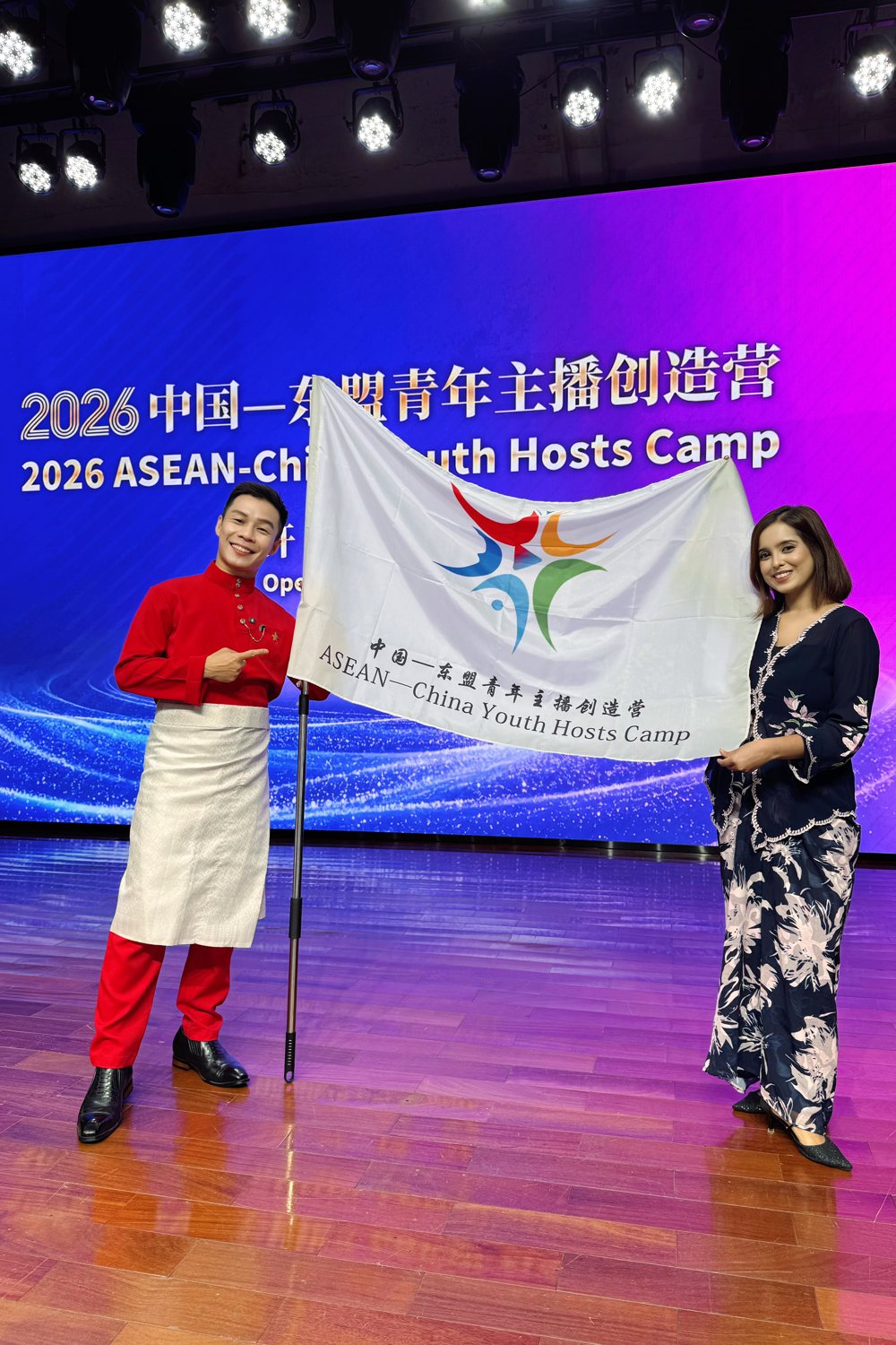 JC 与 Meyra 在开营仪式舞台上，手持2026中国—东盟青年主播创造营旗帜